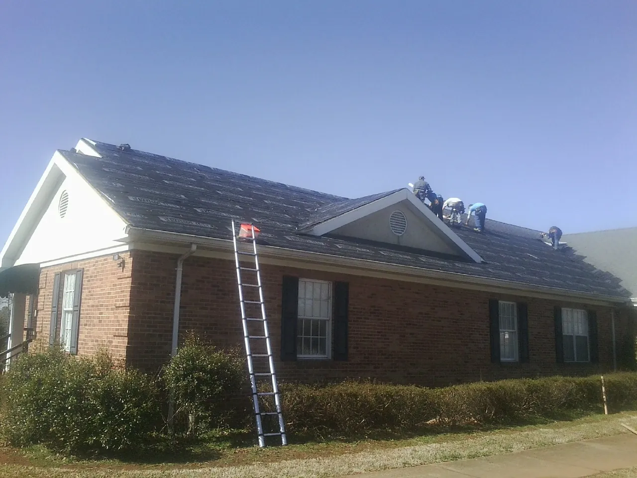 Crew tearing shingles off a brick ranch roof with a ladder set — Emergency Roof Repair work by our crew in Little Cedar