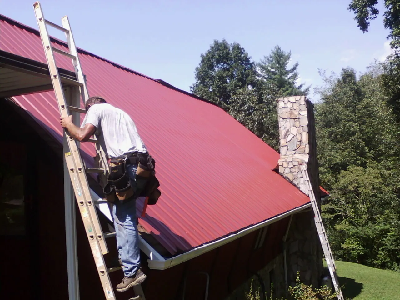 Roofer on a ladder beside a red metal roof and stone chimney — Soffit & Fascia Repair by Roofing Brothers in Little Cedar